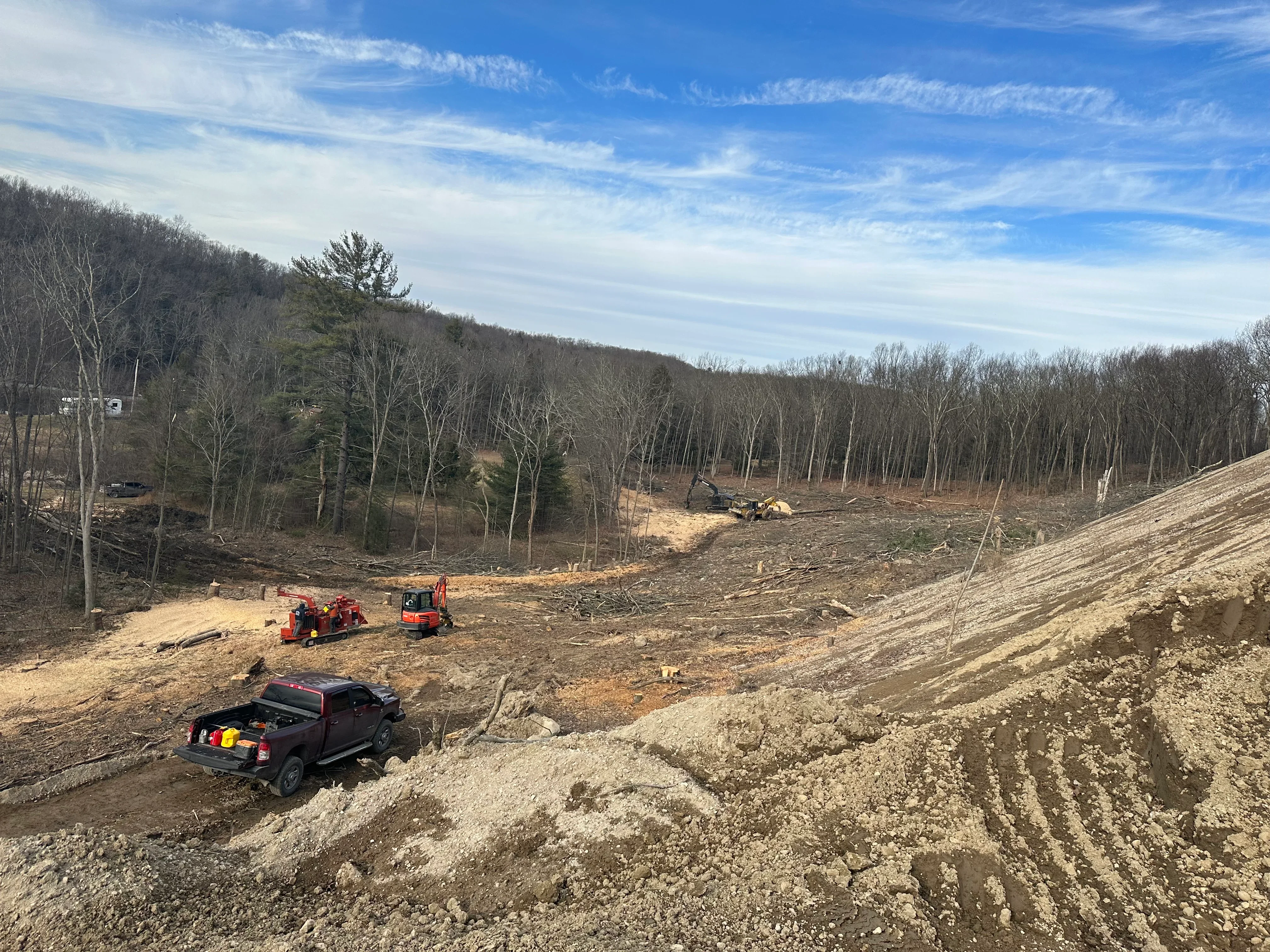 Timber harvest site with red wood chipper, excavator, and cleared hillside surrounded by standing forest