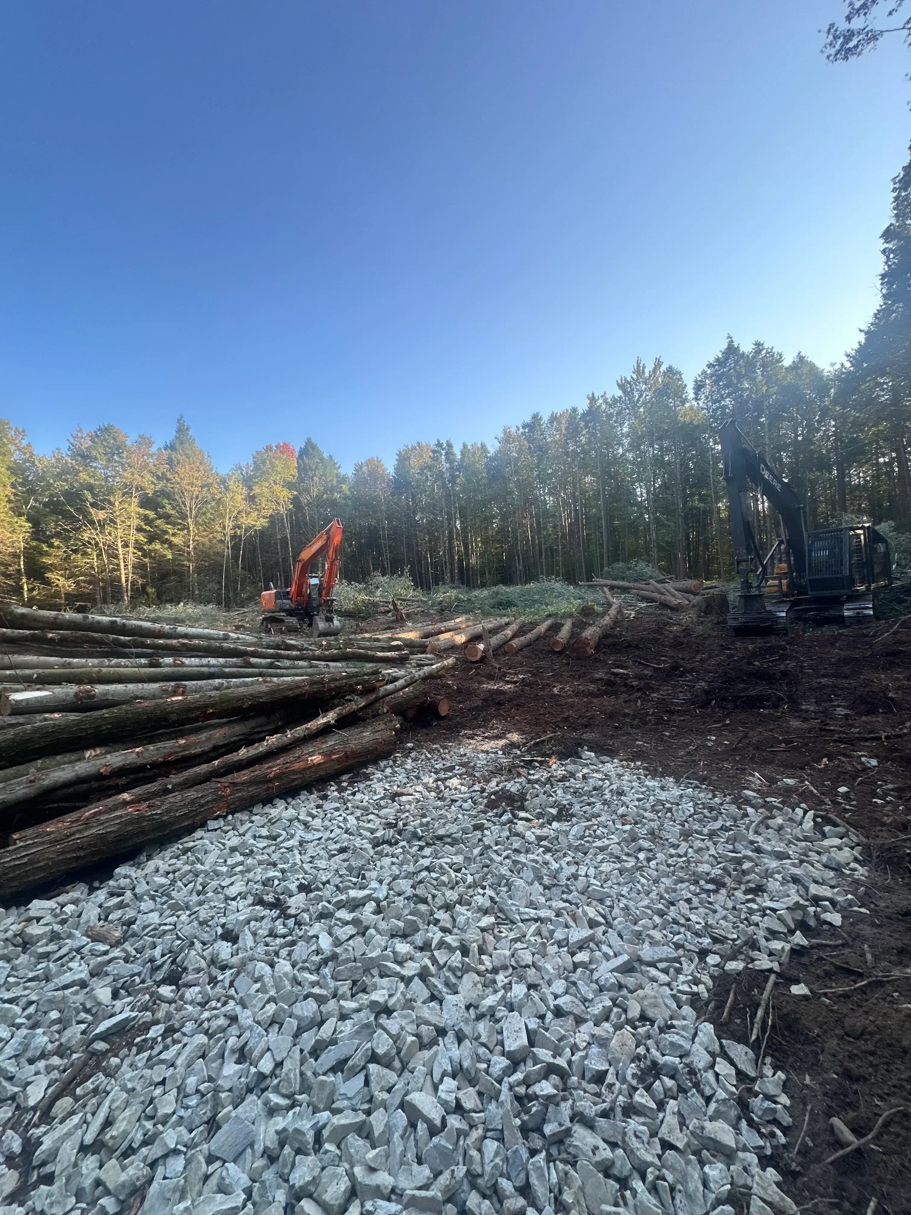 Felled logs and crushed stone aggregate at timber harvest site with equipment and forest backdrop